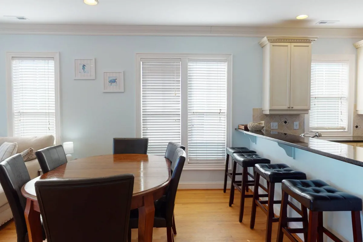 This image features a welcoming interior dining area with a dining table, bar stools, kitchen counter, and large windows. The room exudes warmth and comfort, suggesting a space where family and pets can gather and relax after a fun day at the beach.