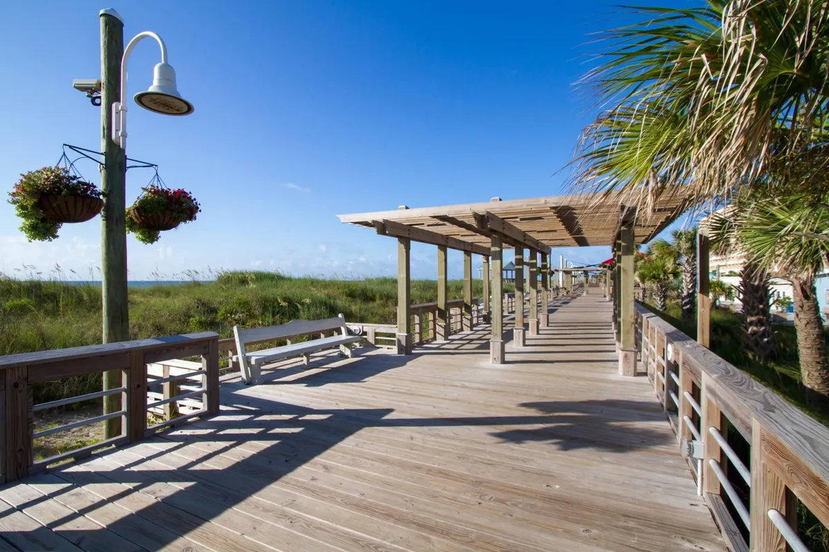 A tranquil wooden boardwalk lined with hanging flower baskets and palm trees leads toward lush surroundings. The scene suggests a pleasant, walkable route to local attractions, encouraging a sense of ease and relaxation.