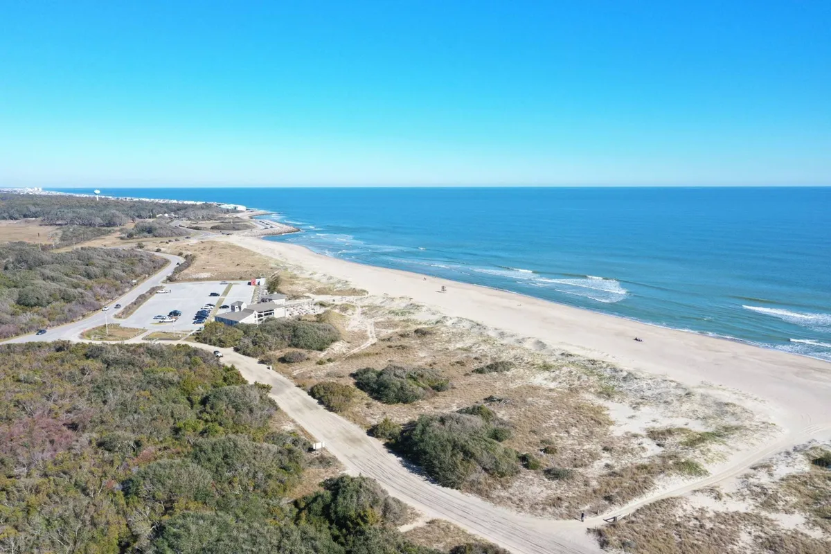 A sunlit, sandy beach unfolds toward a gentle ocean, with grassy dunes in the foreground and a small parking area visible. People enjoy walking, sunbathing, and swimming. The scene is peaceful, evoking a relaxed vacation atmosphere and showcasing direct beach access, parking convenience, and the natural beauty of the area.