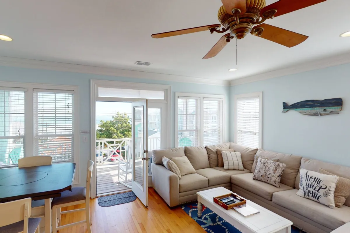 A cozy, sun-drenched open-plan living area featuring a plush sectional sofa, a large wooden dining table, and an open door leading to a balcony. Decorative fish art on the wall ties in subtle coastal touches. Ample windows flood the space with natural light, and the scene evokes a welcoming, communal atmosphere ideal for families and friends to gather, dine, or relax together.