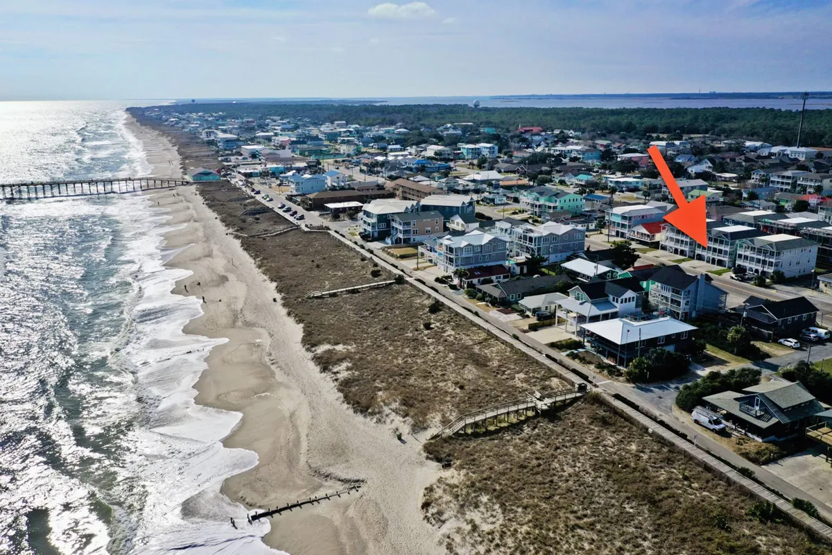An inviting perspective of the beach and ocean, with houses and the pier along the shoreline, capturing the community atmosphere and the natural beauty of Kure Beach. The scene shows gentle waves, sandy stretches, and the potential for multiple beach activities.