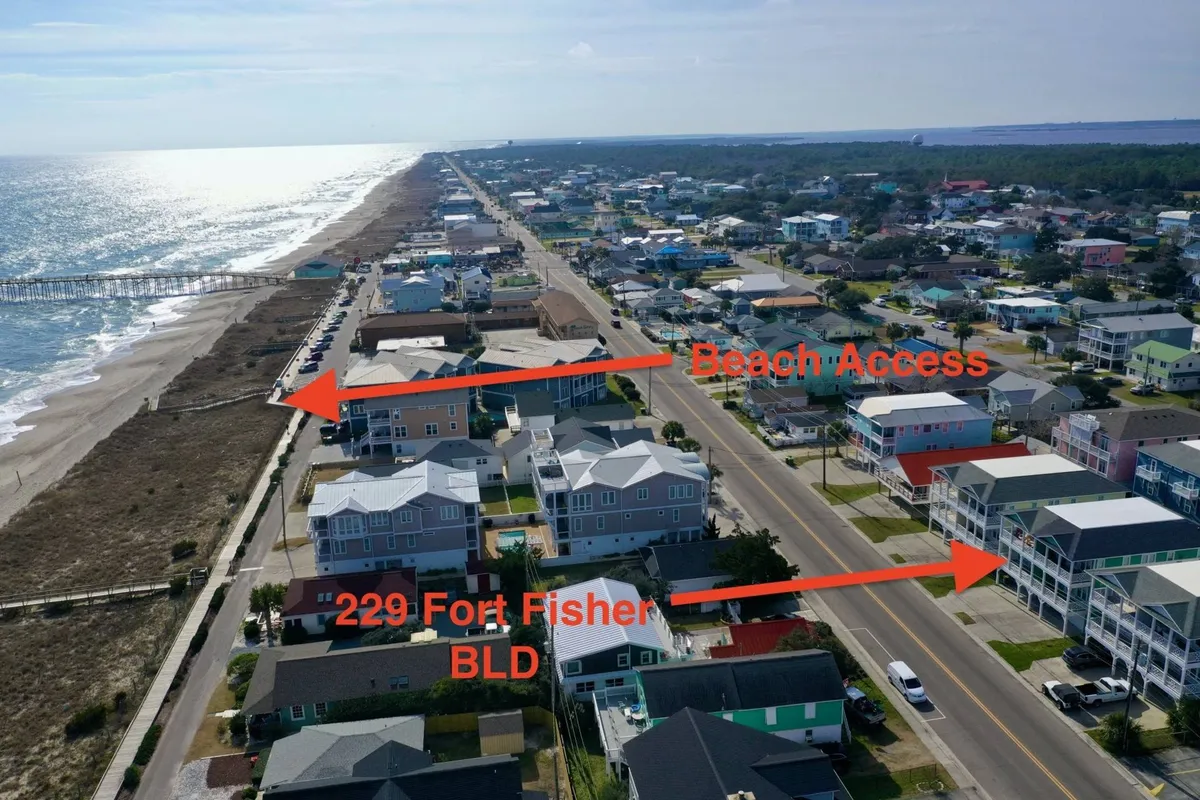 A view of the direct beach access at 229 Fort Fisher Blvd, with sandy paths leading to the shore, neighboring houses visible, and the pier in the distance. The image evokes the feeling of proximity and ease of reaching the ocean from the property.