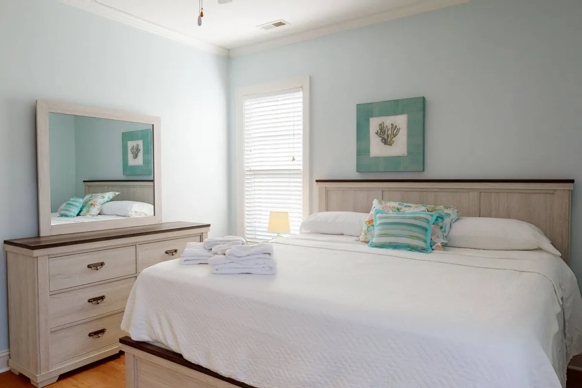 A tranquil bedroom with a bed adorned with decorative pillows, a dresser with a mirror, and coral-themed wall art. Sunlight softly illuminates the space, creating an inviting atmosphere for a restful night's sleep.