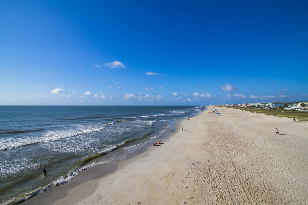 A sun-drenched sandy beach stretches along the shoreline, with people enjoying the waves, some surfing, swimming, and walking under a clear blue sky. The lively atmosphere suggests a hub for water sports and outdoor activities.