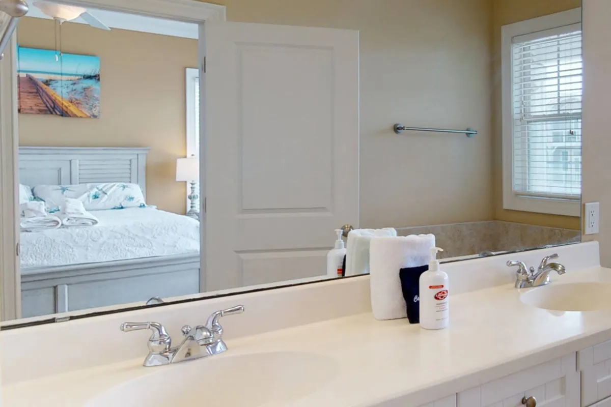 A bright, spacious bathroom featuring a double sink, large mirrored vanity, and a glimpse of an adjacent bedroom. Natural light fills the space, and the decor is fresh and inviting, indicating comfort and convenience for guest routines.