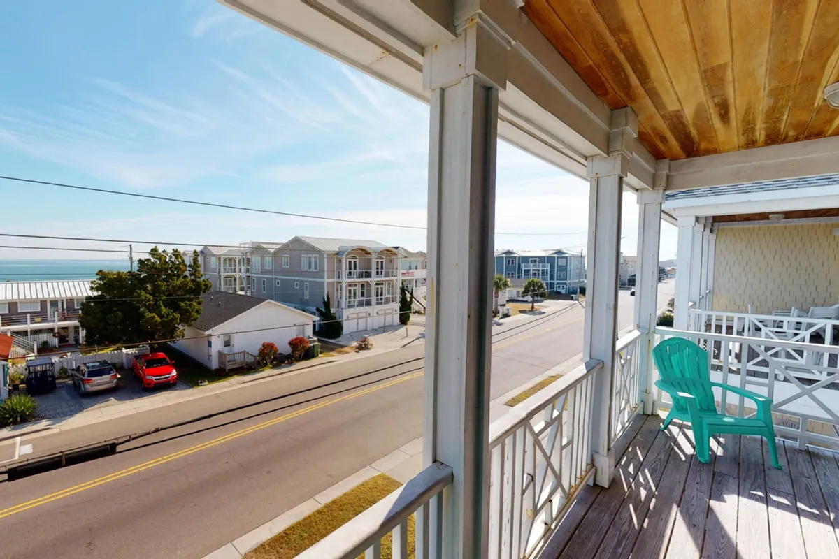 A tranquil balcony scene with a green chair facing the ocean, neighboring houses visible nearby. The setting is perfect for relaxing after a day of adventure, socializing, or simply soaking in the views.
