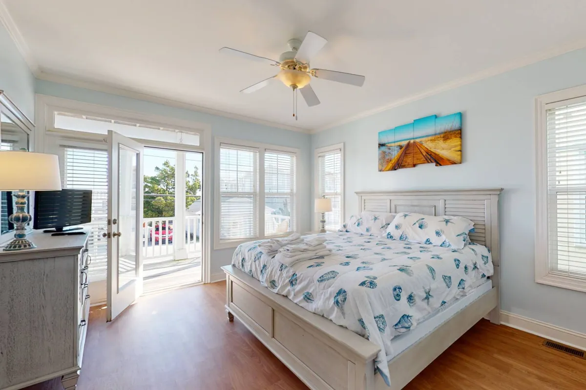 A spacious bedroom featuring a comfortable bed with sunlight streaming in through open doors that lead to a private balcony. The room is tastefully decorated with coastal artwork and includes a dresser and a soft lamp, creating a tranquil retreat. The balcony offers a hint of ocean views, reinforcing the luxury of the master suite.