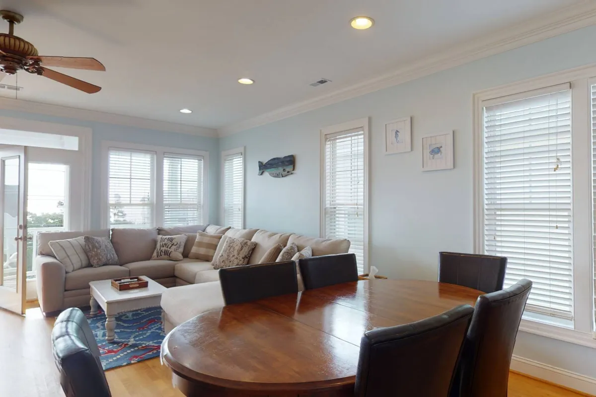 A welcoming living and dining space with a comfortable sectional sofa, eye-catching fish art, a large table set for gatherings, and expansive windows letting in sunlight. The television is subtly visible, reinforcing the space’s suitability for movie nights or group relaxation, all framed by an airy, coastal vibe.