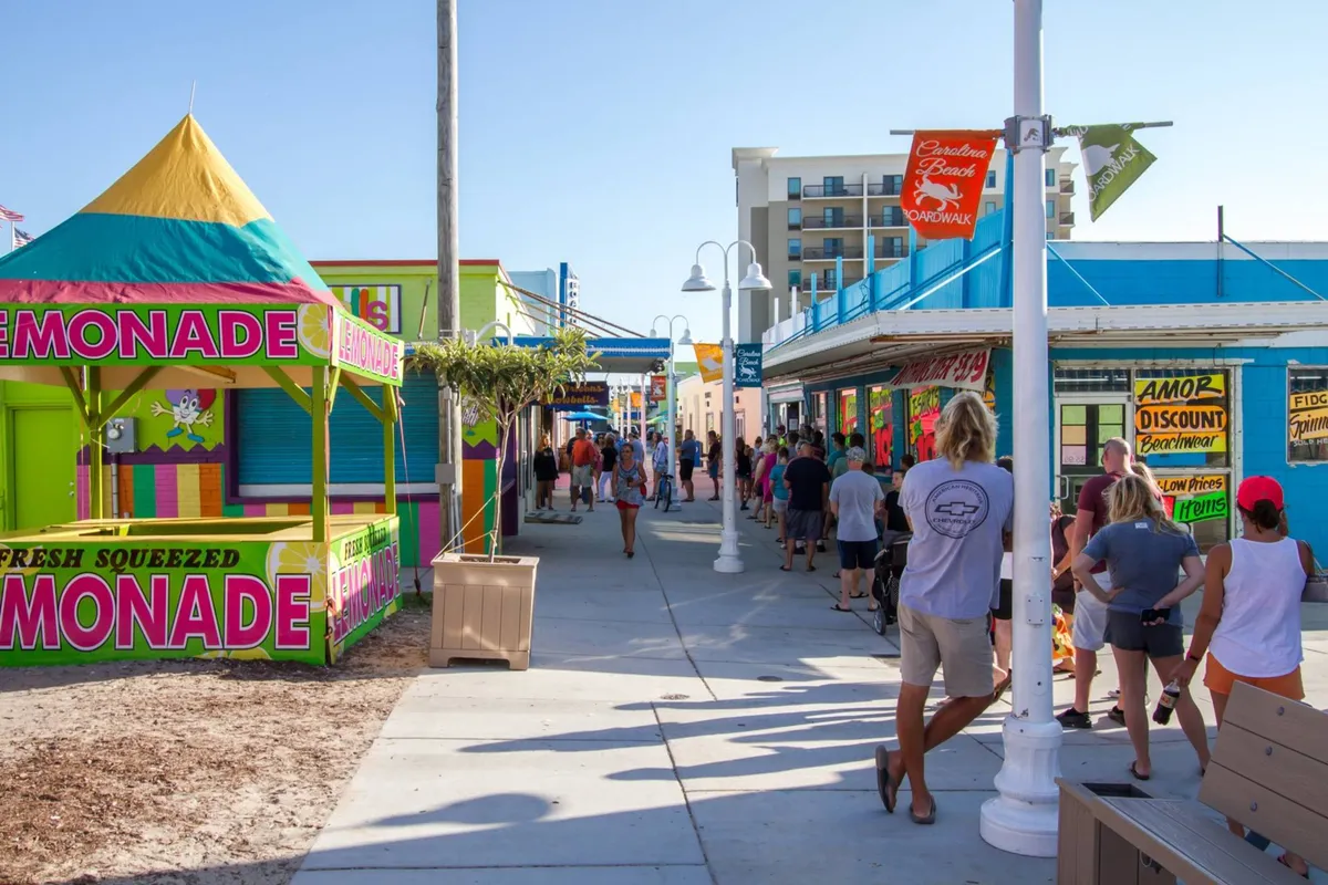 A festive street scene with a colorful lemonade stand, a lively crowd of people enjoying themselves, and bright storefronts that suggest local boutiques and restaurants. The image evokes a vibrant, social atmosphere typical of a walkable beach town.