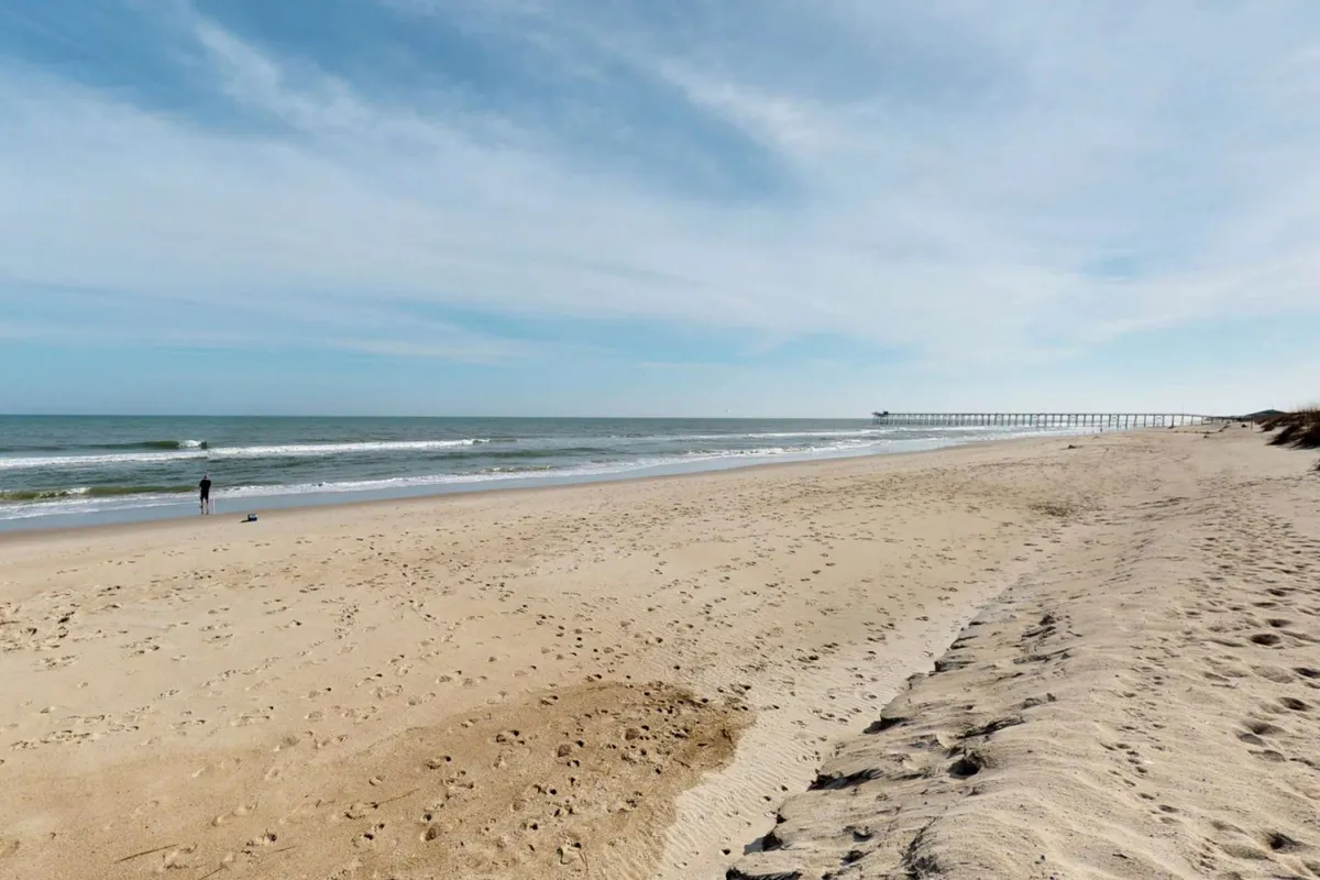 An inviting exterior view capturing a sandy beach with gentle ocean waves and the pier visible in the distance. A solitary figure walks along the shore, evoking a sense of leisurely exploration and freedom.