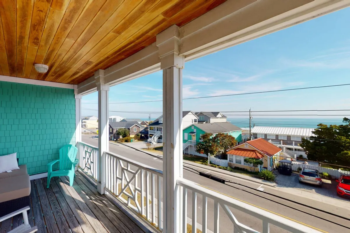 The hero image features the turquoise-walled exterior of Pirate Queens, with a spacious wooden-ceiling porch overlooking the sparkling ocean. The balconies offer sweeping views, inviting guests to relax, socialize, and soak in the tranquil seaside atmosphere just steps from the sand.