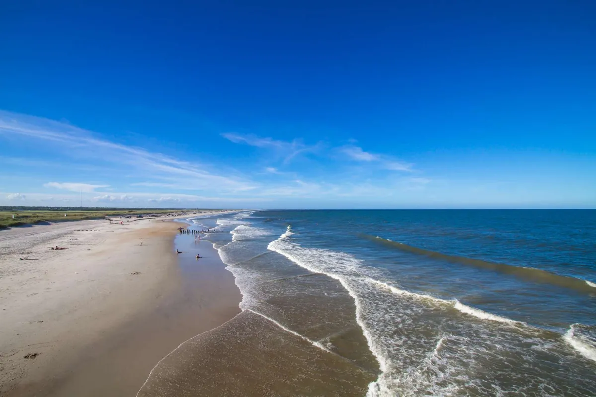 A serene stretch of sandy beach with ocean waves rolling in, a clear blue sky above, and a few distant figures enjoying a walk along the shore. The scene evokes relaxation and possibilities for family fun, such as building sandcastles or taking leisurely strolls.