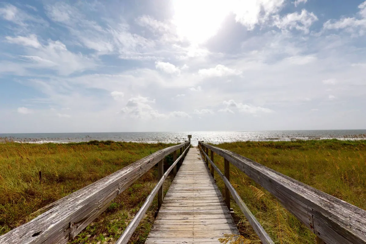 A wooden walkway leads directly toward the ocean, with a wide, sandy beach and the distant horizon under a picturesque sky. The scene is tranquil, evoking the feeling of an early morning stroll or jog along the shore, perfect for dog owners seeking scenic outdoor activities with their pets.