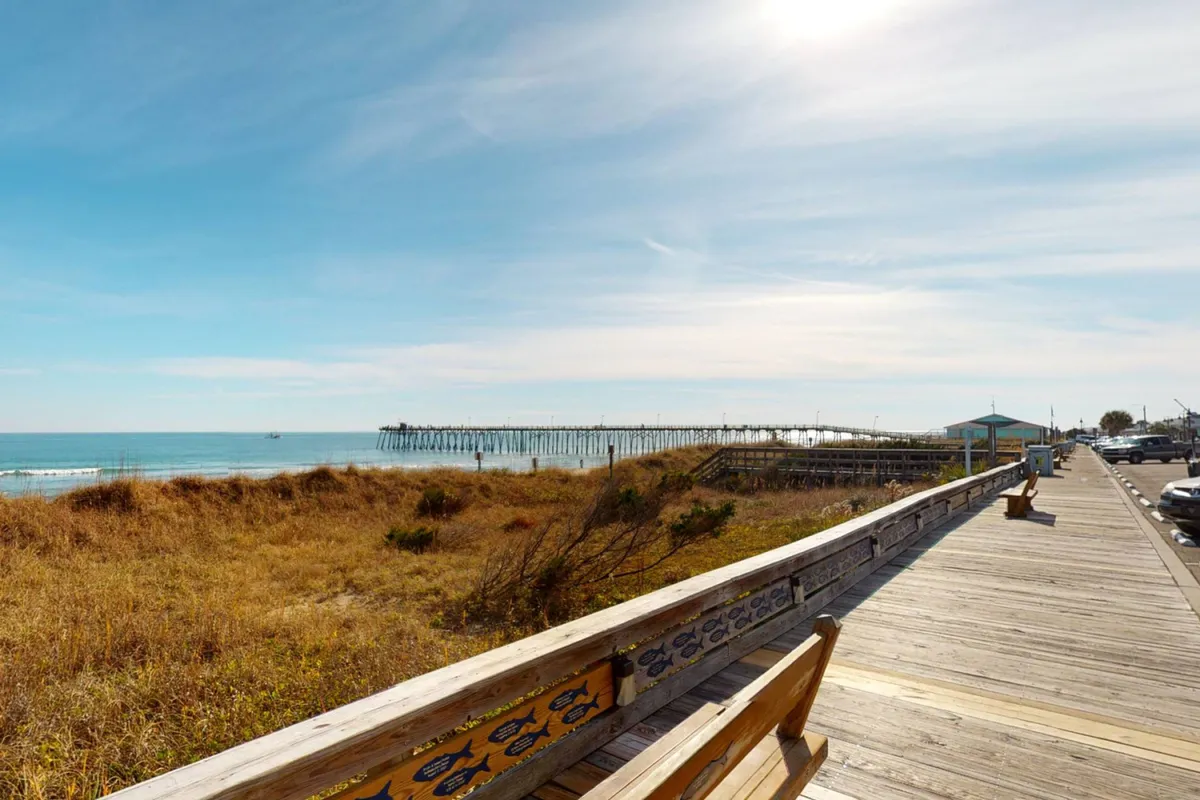 A serene exterior view of the Kure Beach Pier stretching out into the ocean, with the sandy beach and boardwalk in the foreground. The composition showcases the iconic pier, beachgoers strolling along the boardwalk, and a picturesque ocean backdrop.